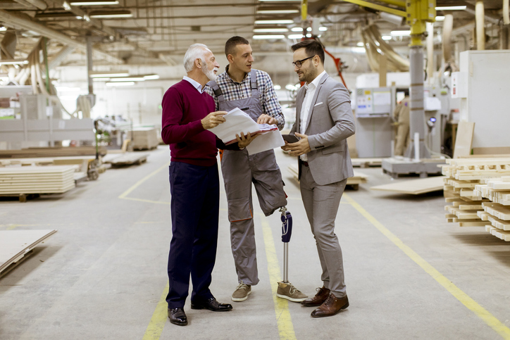 Three men standing and discuss in furniture factory. One man is older, and one of the younger men has limb difference and has a prosthetic leg